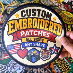 May include: A round, cream-colored embroidered patch with a black border. It features the text CUSTOM EMBROIDERED PATCHES, ALL SIZES, and ANY SHAPE. Graphics include a US flag, a red heart, a mountain landscape, a tiger face, and a star.