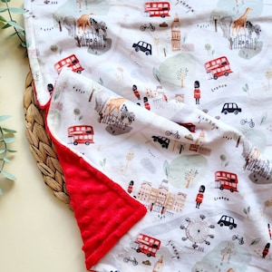 May include: A white baby blanket with a London theme. The blanket features red double-decker buses, black taxis, the London Eye, Big Ben, and the words "London" and "Zoo". The blanket has a red fleece backing.