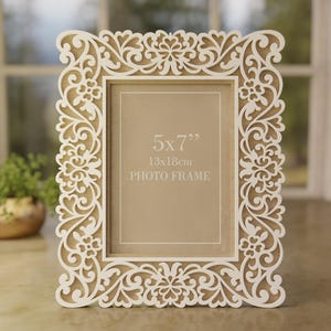 May include: White ornate photo frame with a 5x7 inch opening. The frame has a delicate floral design and the text "5x7" "13x18cm" "PHOTO FRAME" is printed on the front.