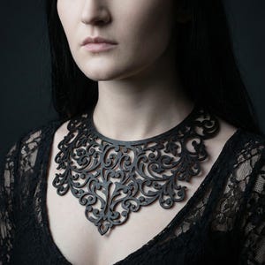 May include: A black laser-cut necklace template with an intricate, symmetrical floral design. The necklace is worn by a person wearing a black lace top. The text "Laser Cut Necklace Template" is at the top.