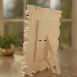 May include: A light-colored wooden picture frame with a decorative, layered edge. The frame has a built-in easel back for tabletop display. The frame is designed with a scalloped edge and is suitable for photos or artwork.