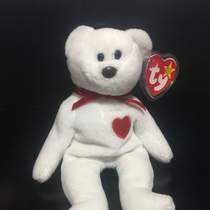 Può includere: Orso di peluche bianco con un cuore rosso sul petto e un nastro rosso intorno al collo. L'orso ha un'etichetta Ty attaccata all'orecchio.