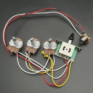 Op de afbeelding: Gitaarbedradingscomponenten, waaronder drie potentiometers, een schakelaar en een jack. De potentiometers hebben bruine bases en zilveren knoppen, verbonden door rode, witte en gele draden. De schakelaar is groen en wit, met een zwarte tuimelschakelaar.