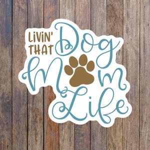 May include: A white sticker with blue and brown text that reads "Livin' That Dog Mom Life" with a brown paw print in the center.