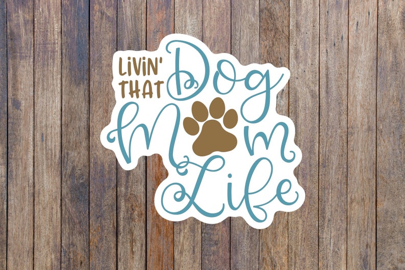 May include: A white sticker with blue and brown text that reads "Livin' That Dog Mom Life" with a brown paw print in the center.