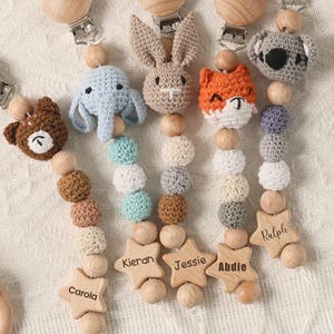 May include: A collection of handmade baby pacifier clips with wooden beads and crochet animal heads, including a bear, elephant, bunny, fox, and koala. Each clip has a wooden star with a name engraved on it. The clips are in various colors.