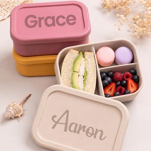 May include: A beige rectangular lunchbox with the name "Aaron" on the lid, open to reveal a sandwich, macarons, and berries. A pink lunchbox with "Grace" on the lid is stacked on top of a yellow one.