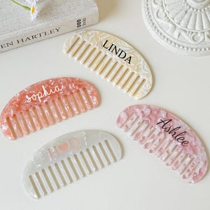 May include: Four personalized hair combs in various colors: cream, pink, and white. Each comb has a name printed on it: "Linda", "Sophia", "Ashlee", and "H♡D". The combs are arranged on a white surface, with a book and decorative item in the background.