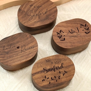 Pode incluir: Quatro caixas de anel de madeira ovais com nomes e datas gravados. As caixas são marrons escuras com grão de madeira visível. O texto inclui "Mr. & Mrs. Sanford" e "Amelia & Raines" com datas. Uma caixa tem "A&J Anna & John".