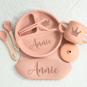 May include: A baby feeding set in pale pink, featuring a plate, bib, cup, spoon, fork, and straws. The set is personalized with the name "Annie" and a crown motif. The plate and bib are designed for easy feeding.