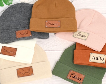 May include: A selection of knitted beanies in a range of colours: grey, tan, pink, orange, and green. Each beanie has a rectangular leather patch with a name. The names on the patches are "Frances", "Pardal", "Jeannie", "Aveal", "Latania", "Edison", and "Aalto".