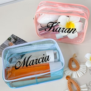 Könnte beinhalten: Zwei transparente Kosmetiktaschen, eine rosa und eine blaue, jeweils mit einem Namen in schwarzer Schrift. Auf der rosa Tasche steht "Karan" und auf der blauen Tasche steht "Marcia". Beide Taschen sind mit Schönheitsprodukten gefüllt. Dekorative Ohrringe und Blumen sind ebenfalls vorhanden.