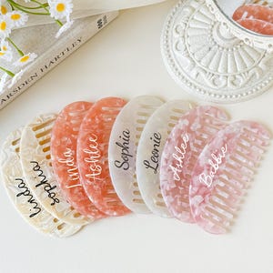 May include: A collection of personalized combs in various colors, including peach, white, and pink, with names like "Linda" and "Ashlee" written on them. The combs have a marbled effect and are arranged on a white surface.