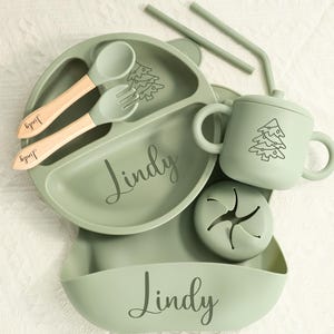 May include: A sage green baby feeding set with the name "Lindy" engraved on the plate, bib, and utensils. The set includes a divided plate, spoon, fork, cup, snack container, and two straws. Each item has a Christmas tree design.