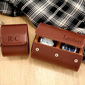 May include: Two brown leather watch cases. One case is closed with the initials "RC" embossed on the front. The other case is open, revealing two watches and the name "Lambert" engraved on the lid. The cases are on a wooden surface.