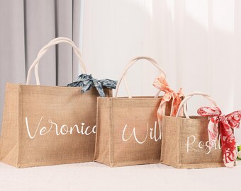 May include: Three burlap tote bags of varying sizes with white handles and personalized names. The largest bag says "Veronica", the middle bag says "Will", and the smallest bag says "Rosie". Each bag has a decorative ribbon.