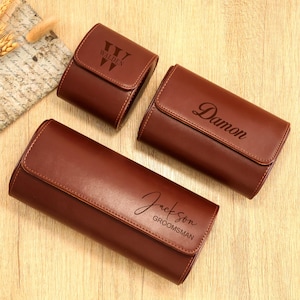 May include: Three brown leather watch cases with personalized text. One case reads "Walden", another "Damon", and the third "Jackson GROOMSMAN". The cases have a rectangular shape with a flap closure.