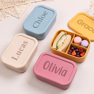 Könnte beinhalten: Eine Sammlung farbenfroher Lunchboxen mit personalisierten Namen. Eine Box ist geöffnet und zeigt ein Sandwich, Macarons und Beeren. Die Boxen sind in Blau, Gelb, Rosa und Beige gehalten, jede mit einem Namen auf dem Deckel.
