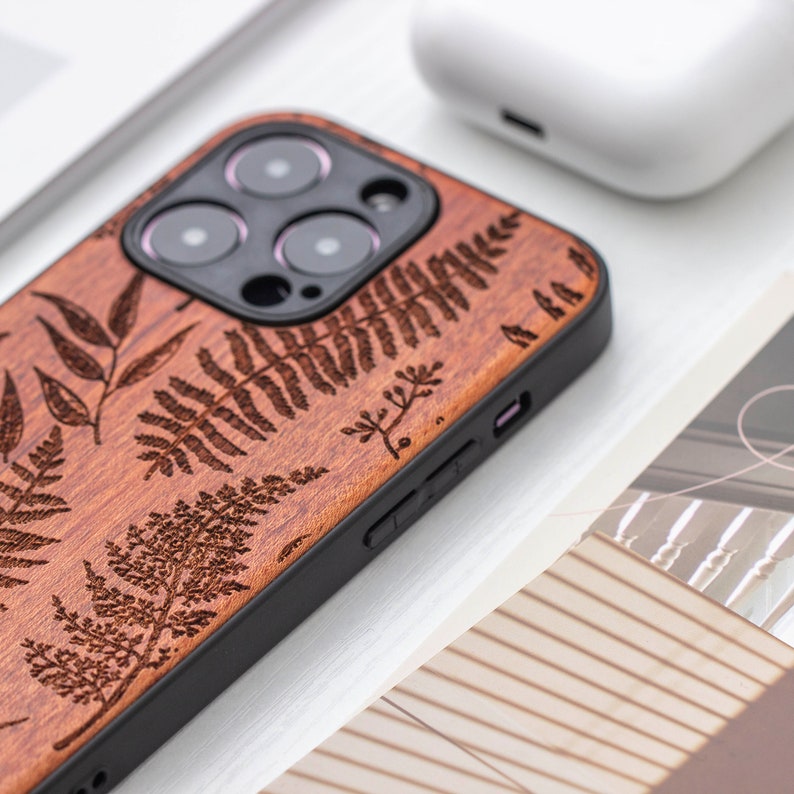 May include: A brown wooden phone case with a laser-etched fern design. The case has a black bumper and a cutout for the camera lens.