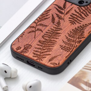 May include: A brown wooden phone case with a black border. The case is decorated with a laser-etched design of ferns and leaves.