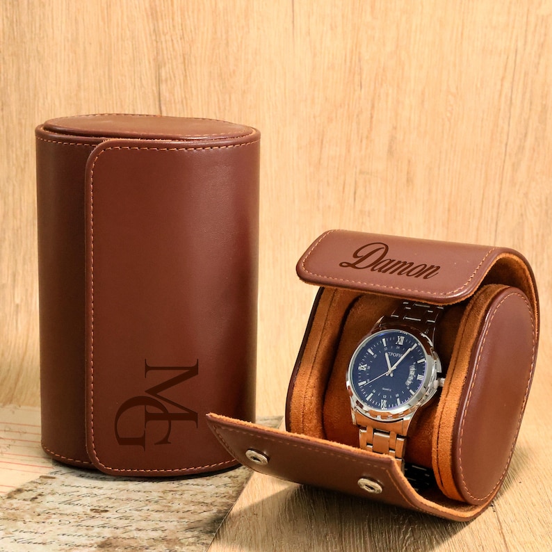 May include: A brown leather watch case with a watch inside. The case is cylindrical with a flap closure. The watch has a blue face and a silver band. The case is embossed with the name "Damon" and initials "MG".