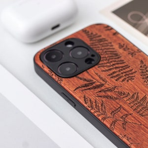 May include: A brown wood phone case with a black border and a fern pattern. The case has a cutout for the camera lens.