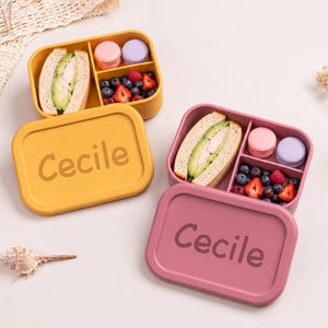 Op de afbeelding: Twee gepersonaliseerde lunchboxen, één gele en één roze, elk met de naam "Cecile" op het deksel. De dozen zijn open en onthullen sandwiches, macarons en verse bessen. De lunchboxen zijn ontworpen met meerdere compartimenten.