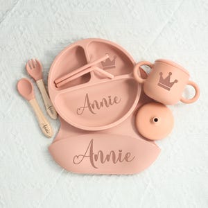 May include: A set of baby feeding essentials in a soft pink hue. The set includes a divided plate, bib, cup with lid, spoon, fork, and straws. Each item is personalized with the name "Annie" and a crown design.