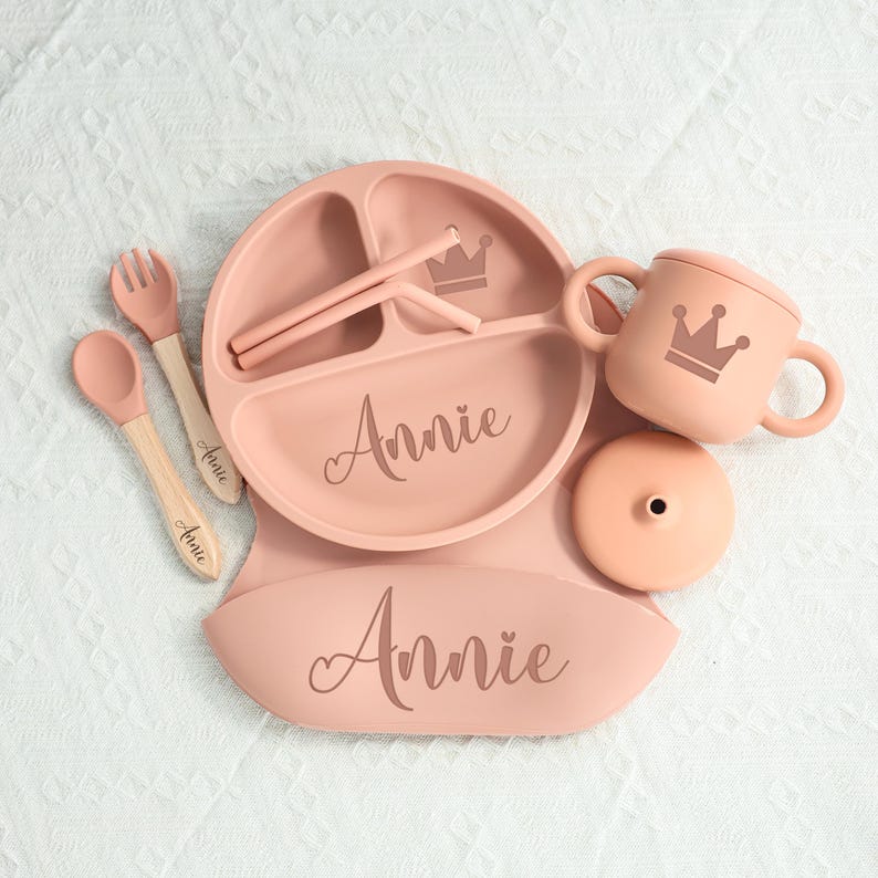 May include: A set of baby feeding essentials in a soft pink hue. The set includes a divided plate, bib, cup with lid, spoon, fork, and straws. Each item is personalized with the name "Annie" and a crown design.