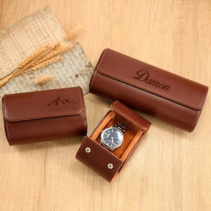 May include: Three brown leather watch cases. One case is open, revealing a silver watch with a black face. Two cases are closed, one with the name "Damon" and the other with the name "Terence" and a mountain graphic.