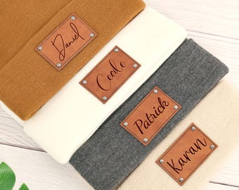 Op de afbeelding: Vier gevouwen mutsen in bruin, wit, grijs en beige tinten. Elke muts heeft een rechthoekige bruine leren patch met een in cursief gegraveerde naam. De patches zijn vastgezet met zilveren klinknagels.