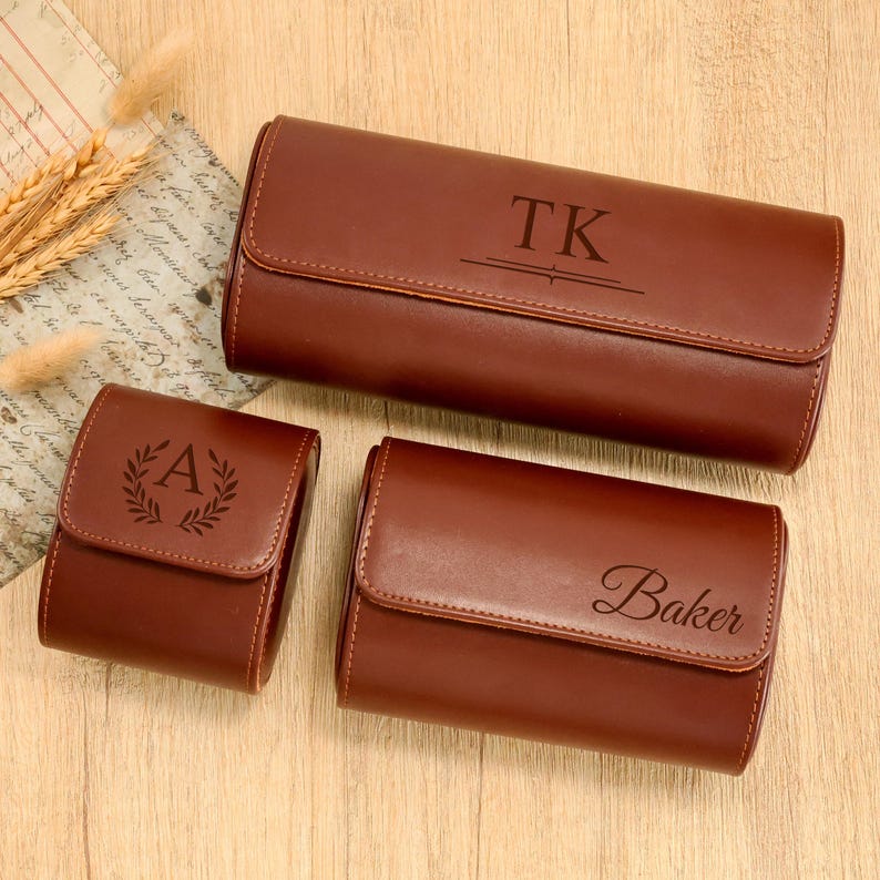 May include: Three brown leather cases with personalized engravings. The largest case has "TK" engraved, the smallest has "A" within a laurel wreath, and the medium case has "Baker" engraved. The cases are on a wooden surface.
