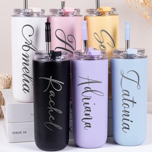 May include: A selection of pastel-coloured personalised tumblers. The tumblers are white, pink, yellow, purple, black, and blue. Each has a name in cursive script. They have lids and straws.