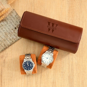 May include: A brown leather watch case with the name "WALDEN" embossed on the front. Two silver watches with metal bands and dark blue and white faces sit on orange velvet cushions. The case is closed and cylindrical.