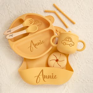 May include: A yellow baby feeding set with a plate, bib, cup, and snack container. The plate has a car design and the name "Annie" is written on the plate and bib. Includes a fork, spoon, and two straws.
