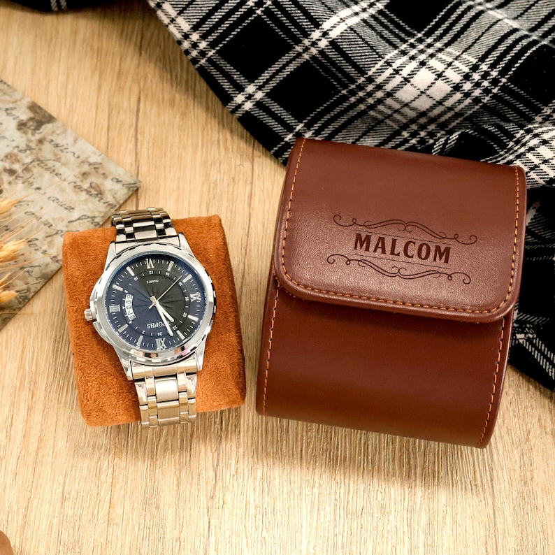 May include: A silver wristwatch with a black dial and a stainless steel band rests on a brown velvet cushion. Beside it is a brown leather watch case with the name "MALCOM" engraved on the front.