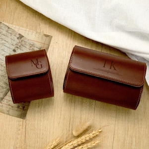 May include: Two brown leather watch cases with personalized initials. The smaller case has "MG" engraved on the flap, while the larger case displays "TK". Both cases are rectangular with a flap closure, set on a wooden surface with a white cloth.