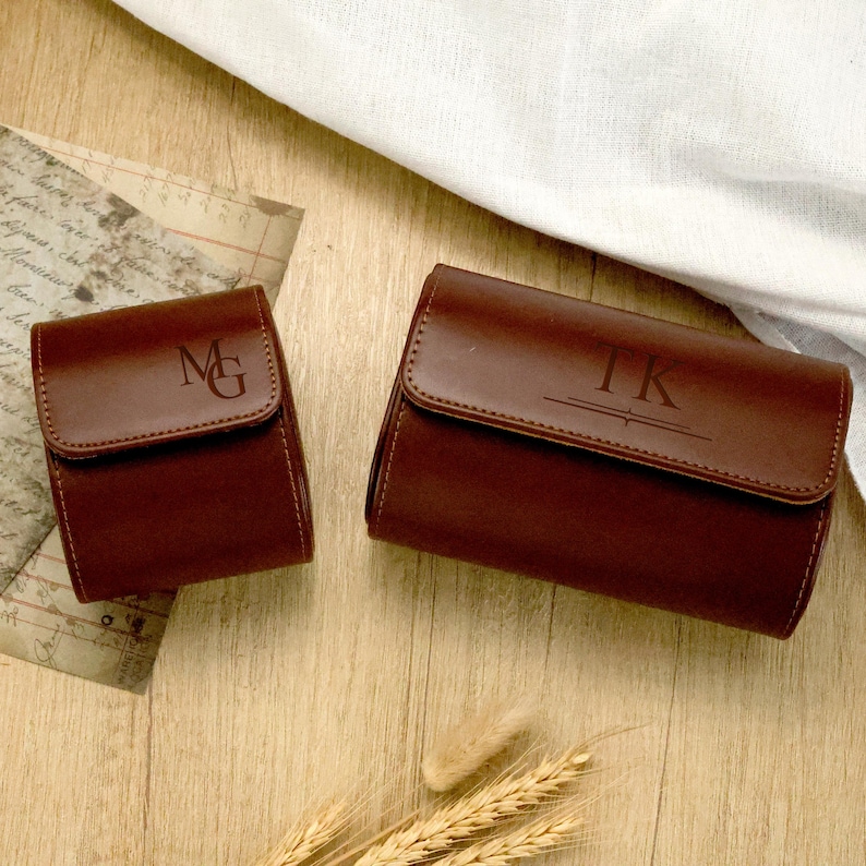 May include: Two brown leather watch cases with personalized initials. The smaller case has "MG" engraved on the flap, while the larger case displays "TK". Both cases are rectangular with a flap closure, set on a wooden surface with a white cloth.