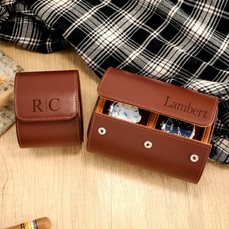 May include: Two brown leather watch cases. One case is closed with the initials "RC" embossed on the front. The other case is open, revealing two watches and the name "Lambert" engraved on the lid. The cases are on a wooden surface.