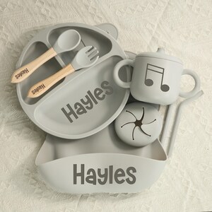 May include: A gray baby feeding set with the name "Hayles" printed on the plate and bib. The set includes a divided plate, spoon, fork, sippy cup with a musical note, and a snack container. The spoon and fork have wooden handles.