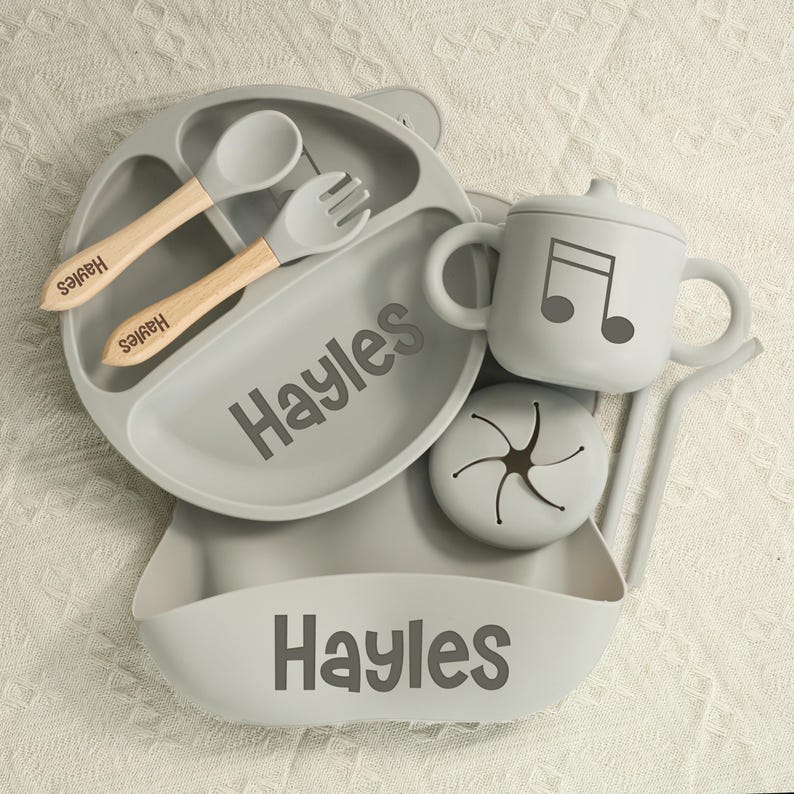 May include: A gray baby feeding set with the name "Hayles" printed on the plate and bib. The set includes a divided plate, spoon, fork, sippy cup with a musical note, and a snack container. The spoon and fork have wooden handles.