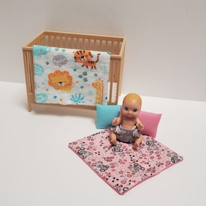 May include: A doll wearing a grey and white outfit sits on a pink and white patterned play mat. A wooden cot with a white and blue animal print blanket is in the background.