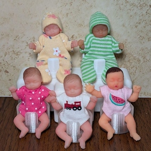 May include: Five miniature baby dolls in a white plastic stand. Two dolls wear hats and long-sleeved outfits in yellow with floral patterns and green stripes. Three dolls wear short-sleeved onesies in pink with white dots, white with a red train, and pink with a watermelon slice.