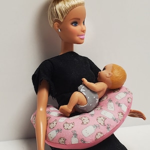K&ouml;nnte beinhalten: Eine blonde Puppe, die ein schwarzes Shirt tr&auml;gt und eine Baby-Puppe h&auml;lt. Die Baby-Puppe sitzt auf einem rosa-wei&szlig;en Stillkissen mit einem Milchflaschen- und Keksmuster.