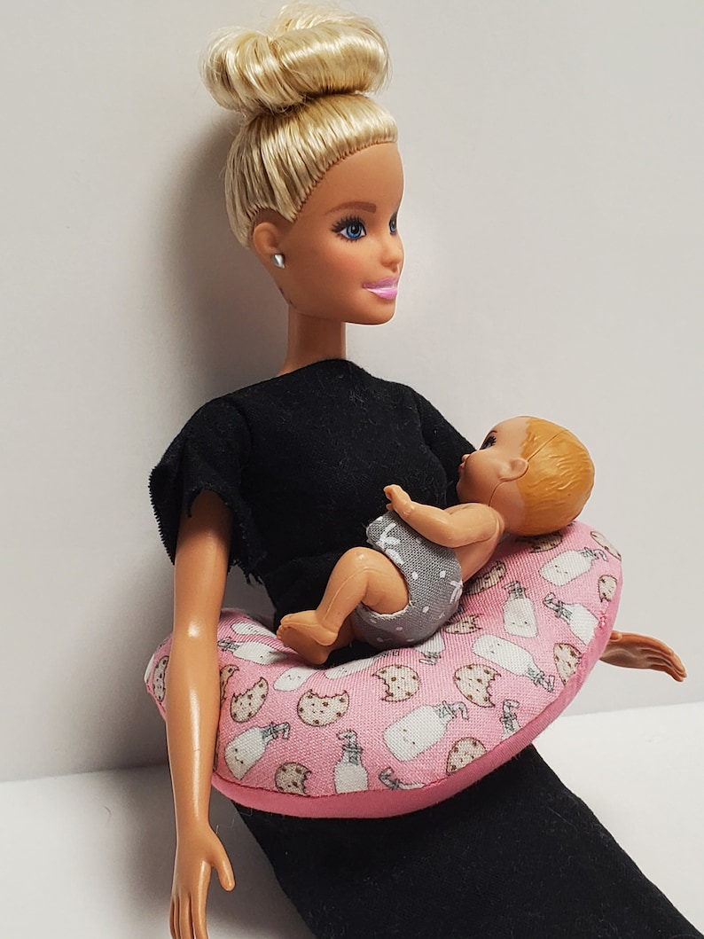 K&ouml;nnte beinhalten: Eine blonde Puppe, die ein schwarzes Shirt tr&auml;gt und eine Baby-Puppe h&auml;lt. Die Baby-Puppe sitzt auf einem rosa-wei&szlig;en Stillkissen mit einem Milchflaschen- und Keksmuster.