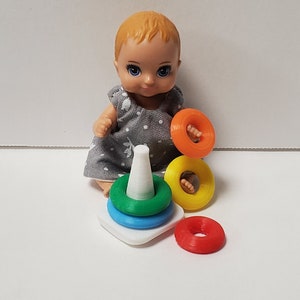 May include: A blonde doll with blue eyes is playing with a colorful stacking toy. The toy has a white cone base and four rings in red, yellow, blue, and green.