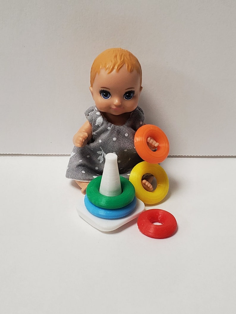 May include: A blonde doll with blue eyes is playing with a colorful stacking toy. The toy has a white cone base and four rings in red, yellow, blue, and green.
