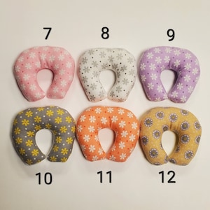 K&ouml;nnte beinhalten: Sechs U-f&ouml;rmige Nackenst&uuml;tzkissen mit verschiedenen Blumenmustern. Die Kissen sind in zwei Reihen mit jeweils drei Kissen angeordnet. Die obere Reihe hat rosa, graue und lila Kissen. Die untere Reihe hat graue, orange und gelbe Kissen. Jedes Kissen hat eine Nummer von 7 bis 12 aufgedruckt.