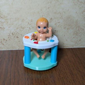 May include: A plastic baby doll with blonde hair and blue eyes sits in a white, blue, and teal baby walker. The walker has a round base and a play area with colourful buttons. The doll is raising its arms.