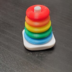 May include: A rainbow-colored stacking toy with a white base. The toy is made of plastic and has six rings that can be stacked on top of each other.